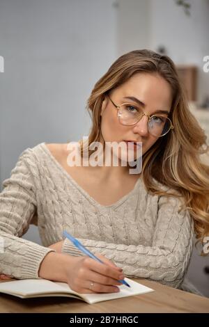 Blonde Frau Blogger in Brille, beige Pullover. In der Küche am Holztisch sitzen, etwas in ihr Notizbuch schreiben und auf Sie schauen. Nahaufnahme Stockfoto