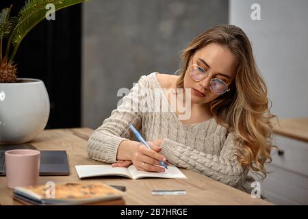 Blonde Model-Blogger in Brille, beigefarbener Pullover. In der Küche am Holztisch sitzen und etwas in ihr Notizbuch schreiben. Nahaufnahme Stockfoto