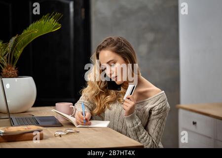 Frau Blogger im beigefarbenen Pullover. In der Küche am Tisch sitzen, mit dem Smartphone sprechen, etwas in das Notebook schreiben und Plastikkarte halten. Nahaufnahme Stockfoto