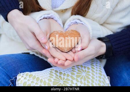 Die Hände der Männer halten die Handflächen der Frauen, in denen Kekse in Form eines Herzens liegen. Ein Paar in blauer Jeans, die einen Lebkuchen in Form eines Herzens hält. Schließen Stockfoto