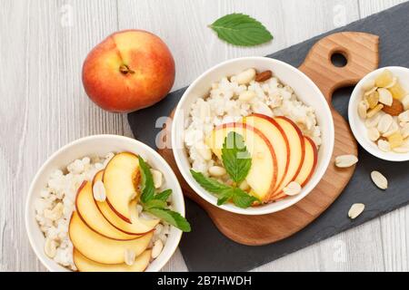 Sorghumporridge mit Pfirsichstückchen, Cashewnüssen und Mandeln in Porzellanschüsseln, frischen Pfirsichen auf Holz- und Steintafeln. Veganes glutenfreies Sorghum Stockfoto