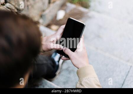 Rückansicht eines hispanischen jungen Mannes, der im Freien mit dem Handy sitzt Stockfoto