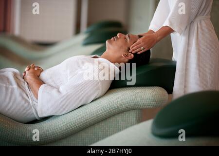 Der Mann im bademantel liegt wieder in einer Spa-Chaiselounge, als eine Frau seinen Kopf massiert. Stockfoto