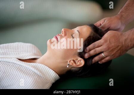 Frau in einem bademantel liegt wieder in einer Chaiselounge, als ein Mann ihren Kopf massiert. Stockfoto