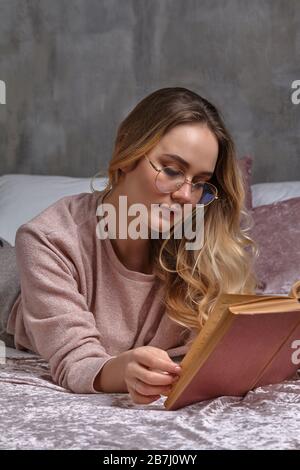 Blondes Mädchen in Brille, lässige Kleidung. Sie legt auf Bett und liest Buch im Schlafzimmer. Student, Blogger. Innen mit grauer Wand. Nahaufnahme Stockfoto