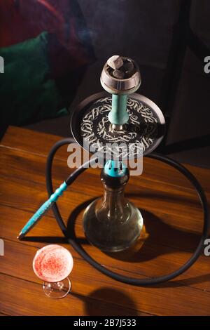 Ein Glas Himbeercocktail neben einem Hookah auf einem Holztisch in einer dunklen Bar. Alkohol- und Rauchkonzept. Stockfoto