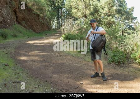 Junge Reisende in einer Kappe mit Rucksackhaltestellen auf der örtlichen Straße halten eine Plastikflasche Wasser und ruht. Kiefernwald in der Nähe des Vulkans Arenas Negr Stockfoto