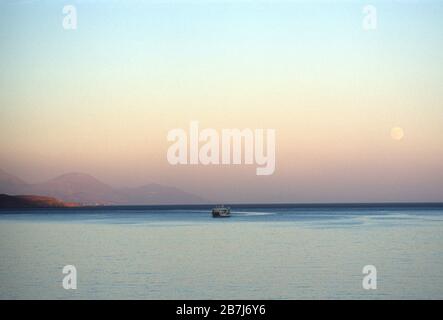 Das Fährschiff Daskalogiannis an einem ruhigen libyschen Meer, das von Chora Sfakia (im Hintergrund gesehen) aus segelt und sich bei Sonnenuntergang dem Dorf Loutro, Crete, Griechenland nähert, mit dem Vollmond am Himmel und den Bergen hinterher. Stockfoto