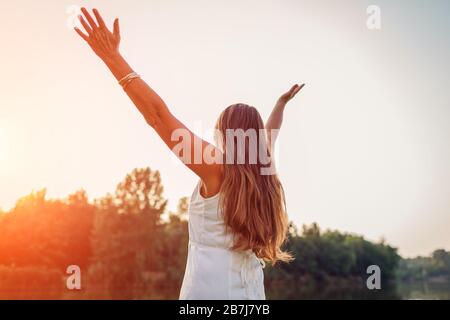 Reife Frau mittleren Alters, die den Sonnenuntergang mit den Armen genießt, die sich im Frühlingspark glücklich fühlen. Seniorin bewundert Landschaft Stockfoto