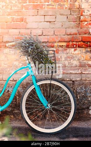 Helles, blaues Vintage-Fahrrad mit Lavendelbouquet im Korb, das gegen die schäbige Ziegelwand auf der Straße platziert ist Stockfoto