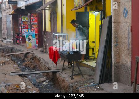 Ein afrikanischer Mann kocht mit einem Rauchgrill über einem offenen Abwassersystem. Großes Loch in der Straße, das schmutzige Wasser strömt. Madagassisches Restaurant in Antananarivo Stockfoto