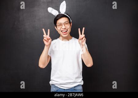 Urlaub, Party und Osterkonzept. Fröhlicher süßer asiatischer Kerl in Kaninchenohren und Gläsern, die Friedenszeichen zeigen, sieht kawaii an, der an einem Tag der orthodoxen Feier feiert Stockfoto