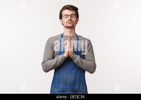 Porträt eines friedlichen und entspannten jungen Mannes in der Schürze, Vorbereitung, Entspannung oder Stress während der Meditation freisetzen, Augen schließen und Hände im Beten halten, p Stockfoto