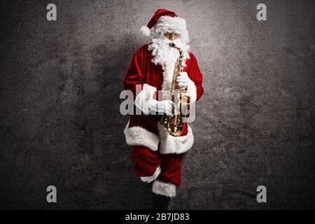 Der Weihnachtsmann spielt ein Saxofon und lehnt sich an eine rostige graue Wand Stockfoto