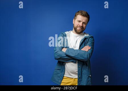 Bescheidener, ruhiger Mann steht mit gekreuzten Armen, Blick auf die Kamera und lächelnd niedlich, mit Kopierraum, auf blauem Hintergrund. Stockfoto