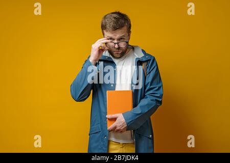 Student oder junger Wissenschaftler in einem Regenmantel und mit einem Buch, hält Brille in der Hand und sieht selbstbewusst aus, auf gelbem Hintergrund. Stockfoto