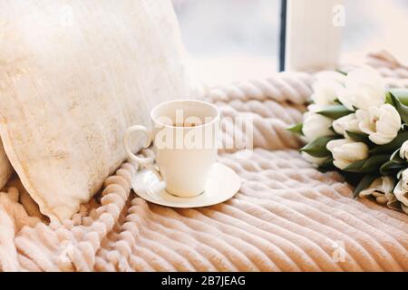 Blumenstrauß aus frischen weißen Tulpen und weißer Tasse Kaffee auf einem gemütlichen Bett mit beigefarbener Plüschdecke und Kissen im hellen Schlafzimmer Stockfoto