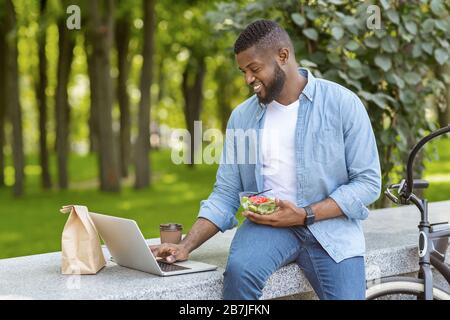 Fröhlicher afroamerikanischer Kerl, der Laptop verwendet und im Freien Mittagessen isst Stockfoto