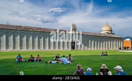 Die Touristen verbringen eine Rast auf dem Rasen der Piazza dei Miracoli vor dem Hintergrund der Außenwand der Camposanto Monumentale, Pisa, Toskana, Stockfoto