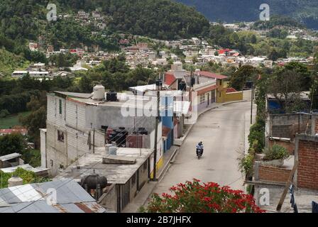 Person auf einem Moped zwischen bunten Häusern mit einem Hintergrund von Bergen und Bäumen in San Christóbal de las Casas, Mexiko Stockfoto