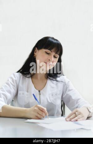 Ein attraktiver Frauenarzt mittleren Alters schreibt ein Rezept aus und macht Notizen auf der Patientenkarte im Büro am Schreibtisch. Spezialist für den täglichen Bereich Stockfoto