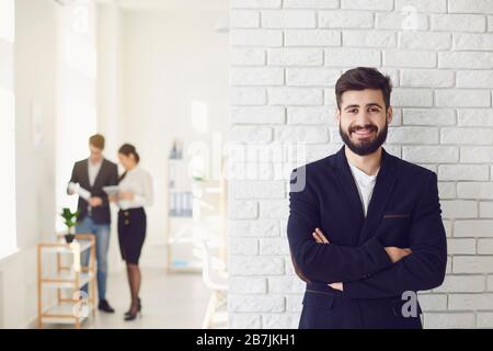 Positiver, glücklicher bärtiger Geschäftsmann in einer Jacke lächelnd, während er vor einem weißen Bürohintergrund steht. Stockfoto