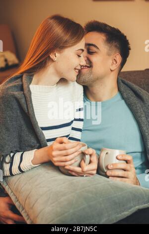 Wunderschönes kaukasisches Paar, das eine Kaffeepause hat, während es auf dem Sofa sitzt und sich küsst Stockfoto