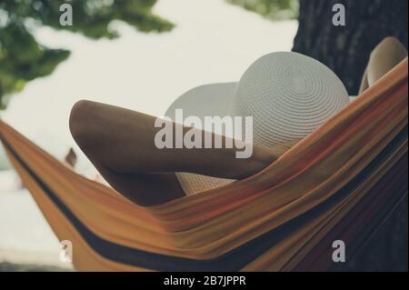 Blick von hinten auf eine Frau mit weißem Strohhalm Sommerhut, die in einer Hängematte mit Blick auf den Ozean genießt. Stockfoto