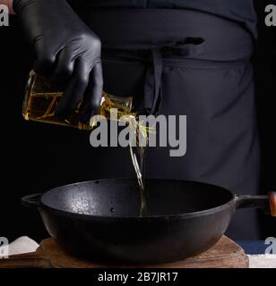 Chefkoch mit schwarzen Latexhandschuhen gießt Olivenöl aus einer transparenten Flasche in eine schwarze gusseiserne Bratpfanne aus der Nähe Stockfoto