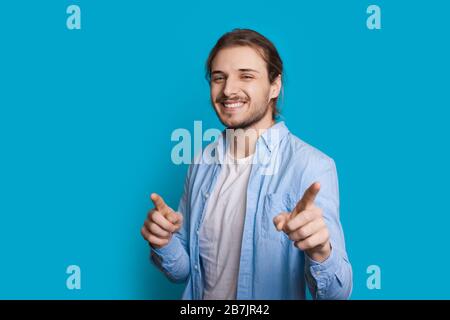 Der gutaussehende kaukasische Mann mit Borsten und langen Haaren zeigt auf die Kamera, während er auf einem blauen Hintergrund lächelt Stockfoto
