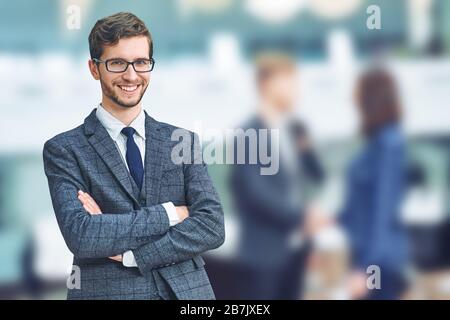 Selbstbewusster und erfolgreicher Geschäftsmann. Fröhlicher junger Kaukasischer in formalwear, die Arme gekreuzt und lächelnd halten, während seine Kollegen darauf stehen Stockfoto