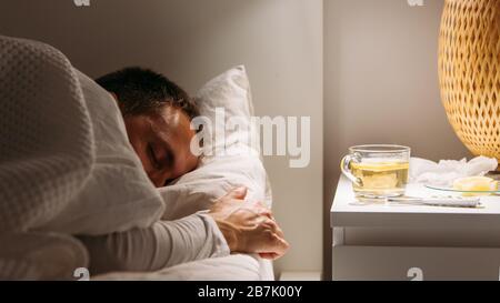 Kranker Mann schläft mit einer Decke bedeckt, die im Bett mit hohem Fieber liegt und im Wohnzimmer ruht. Er ist erschöpft und leidet an Grippe mit einer Tasse Tee Wi Stockfoto