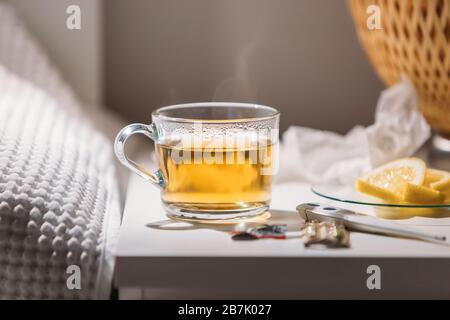 Grippe, Grippesaison, Kaltkonzept. Nahaufnahme der Tasse heißen grünen Tees, Pillen, Digitalthermometer und Untertasse mit Zitronenscheiben am Nachttisch, Stockfoto