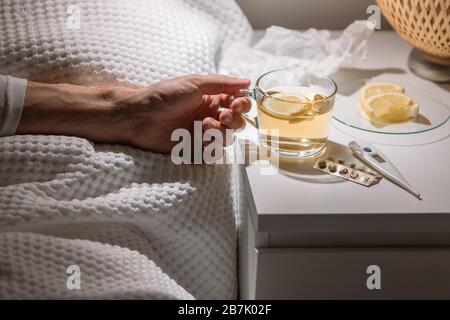 Kranker Mann, der im Bett unter der Decke liegt und eine Tasse heißen Tee mit Zitrone in der Nähe hält. Männer trinken warmes Getränk, um gut von Grippe, Fieber und Virus zu bekommen Stockfoto