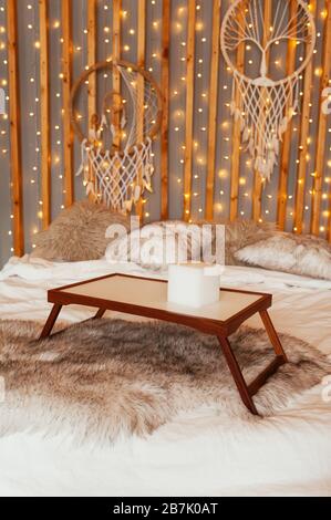 Leerer Holztisch mit Frühstückstisch im Bett. Boho Schlafzimmer, Wanddekor aus Holz mit Girlanden, Lampen und Traumfängern. Stockfoto