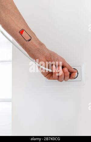 Symbol für niedrigen Batteriestand an Hand/Handgelenk des Menschen. Kreatives Bild müde Mann hält weißen Stecker und lädt sich von der Steckdose. Überarbeit, Stockfoto