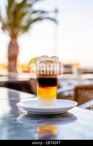 Kaffee kombiniert mit Whisky Condensed Milk Zimt Creme und Zitrone in einer gläsernen Tasse auf einer Terrasse mit Palme im Hintergrund Stockfoto