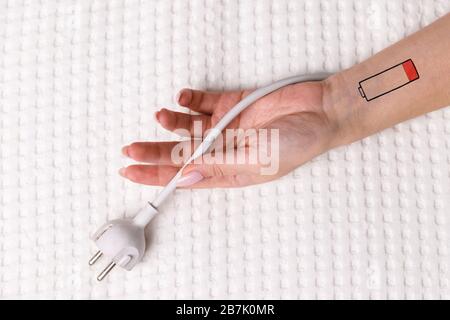 Symbol für niedrigen Batteriestand an Hand/Handgelenk des Menschen. Kreatives Bild einer müden Frau, die auf dem Bett liegt und den Stecker des Kabels/des weißen Steckers hält. Überarbeit, erschöpft, Stockfoto