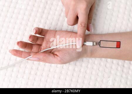 Symbol für niedrigen Batteriestand auf der menschlichen Hand. Müde Frau, die auf dem Bett liegt und versucht, das Gerät über USB aufzuladen und das Kabel mit dem Finger zu halten. Überarbeit, erschöpft, Stockfoto