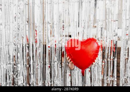 Hände, die einen großen Folienballon in Form eines Herzens auf glattem Hintergrund von silberfarbenen Folienstreifen halten. Bild der Liebe zum Erdtag. Stockfoto
