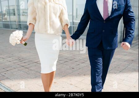 Der Bräutigam in einem blauen Anzug und die Brautpaar in einem stylischen knielangen Kleid und einem beigefarbenen Fellmantel und einem weißen Strauß der Brautpaar halten die Hände und gehen auf den Stockfoto