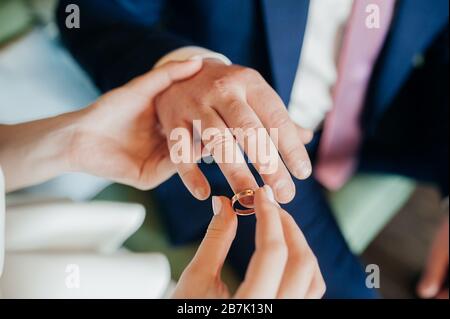 Eine Braute in einem weißen Kleid legt einen goldenen Ring in einem blauen Anzug auf die Hand eines Mannes. Hände der frisch vermählten Nahaufnahme, keine Gesichter. Stockfoto