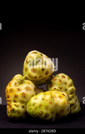 Vertikaler Rahmen von vielen Noni- oder Käsefrüchten, die mit verschiedenen Farbtönen kombiniert sind, die vor dunklem Hintergrund kontrastiert werden Stockfoto