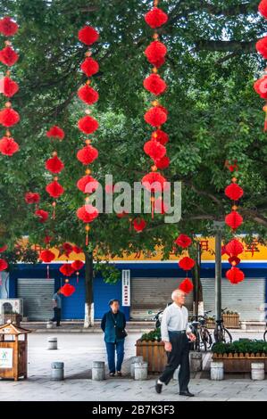 Guilin, China - 11. Mai 2010: Mehrere Ringe von roten Luftballons hängen von Baumzweigen mit grünem Blattwerk. Straßenszene mit Menschen und geschlossenem Laden. Stockfoto