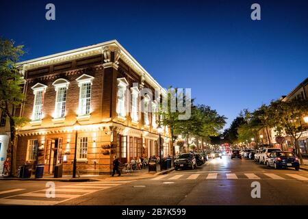 ALEXANDRIA, Virginia, USA – am Ende der King Street in der Altstadt von Alexandria in der Abenddämmerung, wo die historische Hauptstraße auf das Ufer des Potomac River trifft. Die Straße wird von warmen Straßenlaternen beleuchtet, die die charmante Kolonialarchitektur und das geschäftige Treiben der Geschäfte und Restaurants während des Tages zum Abend zeigen. Stockfoto