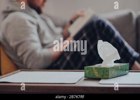 Schachtel mit Geweben auf dem Tisch, Mann auf dem Hintergrund auf einem Sofa sitzend, ein Buch lesend. Konzept "krank zu Hause". Stockfoto