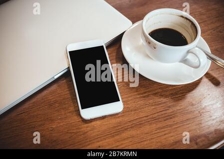 Top-View-Mockup-Bild eines weißen Mobiltelefons mit leerem schwarzem Desktop-Bildschirm auf einem Computerlaptop mit Kaffeetasse Stockfoto