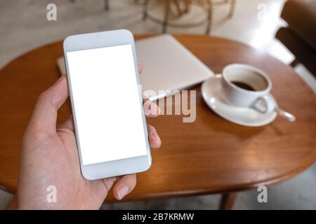 Mockup-Bild des handgehaltenen weißen Mobiltelefons mit leerem Desktop-Bildschirm mit Kaffeetasse und Laptop auf dem Tisch Stockfoto