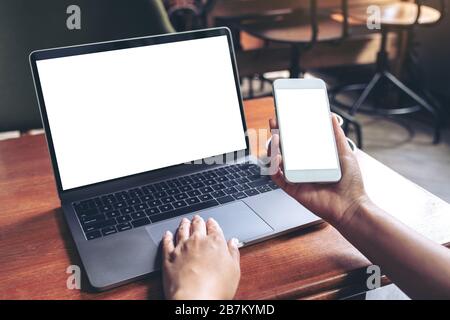 Eine Frau, die ein verspottes Mobiltelefon mit leerem weißen Bildschirm und Laptop auf Holztisch im Café hält und verwendet Stockfoto