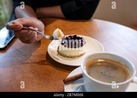 Die Hand einer Frau, die eine Gabel hält, um ein Stück Blaubeerkäse zu schneiden, um mit einer Kaffeetasse auf Holztisch zu essen Stockfoto